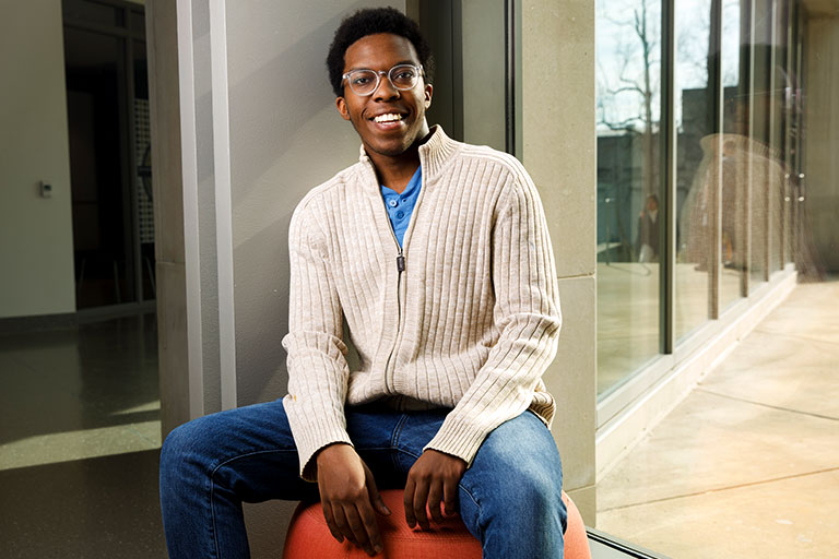 Elvin Irihamye sits on a stool in front of a large window while smiling at the camera.