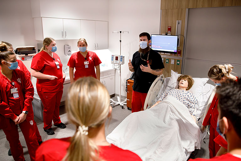 A group of students stands around a simulation mannequin in a hospital room.